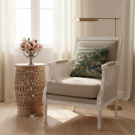 A white woven armchair with beige cushions sits in a bright room with sheer curtains, a carved wooden side table with pink flowers, and a brass floor lamp. A decorative throw pillow rests on the chair.