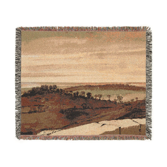 A rectangular woven throw blanket with fringed edges, featuring a scenic landscape print in muted earth tones of beige, brown, and gray.