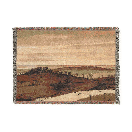 A rectangular woven throw blanket with fringed edges, featuring a landscape scene in muted earth tones of beige, brown, and gray.