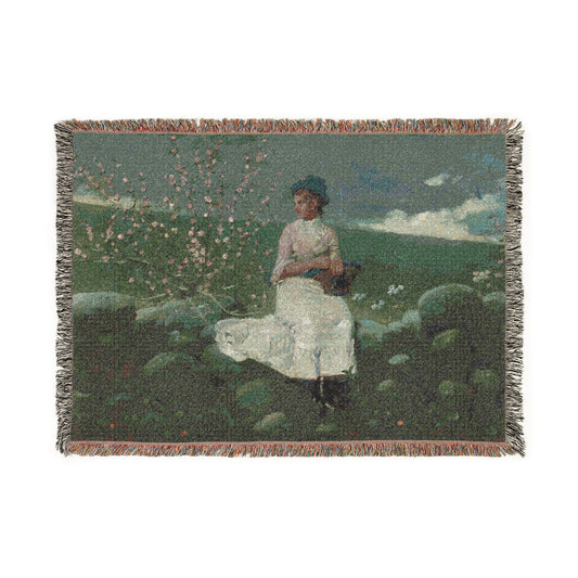 A rectangular woven throw blanket with fringed edges, featuring a vintage-style painting of a woman in a white dress sitting outdoors among green hills, rocks, and pink flowers under a blue sky.
