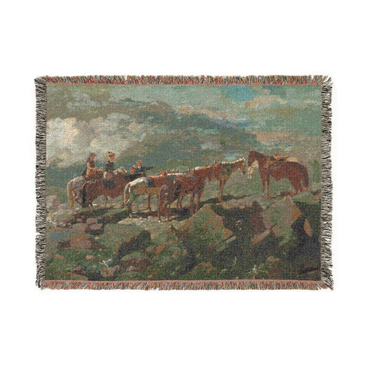 A woven throw blanket featuring a scenic landscape print of riders on horseback with a herd of horses on a rocky hillside, finished with fringed edges.