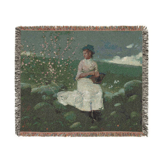 A woven throw blanket featuring a vintage-style painting of a woman in a white dress sitting outdoors among flowering bushes, with fringed edges.