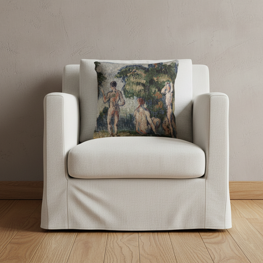 A square throw pillow featuring an impressionist painting of three nude figures in a lush green landscape, styled on the back of a cream-colored armchair against a textured beige wall and light wood flooring.