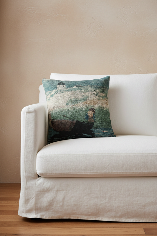 A square throw pillow featuring a textured, vintage-style painting of a lighthouse and a rowboat on the water, styled on a white fabric slipcovered sofa against a beige wall.