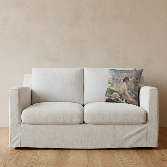 An off-white slipcovered loveseat sofa with a textured fabric, set against a warm beige plaster wall and light wood flooring. A square throw pillow featuring a colorful impressionist painting of a woman is placed on the right side of the sofa.