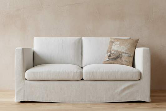 A light beige slipcovered loveseat sofa with two seat cushions and two back cushions, styled with a textured brown and beige throw pillow, set against a warm beige plaster wall and light wood floor.