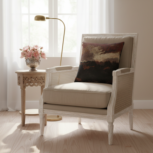 A white-washed wicker armchair with beige cushions sits in a bright room beside a small carved side table with pink flowers and a floor lamp. A dark abstract-patterned throw pillow rests on the chair.