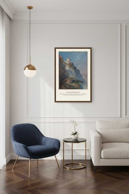 A framed art print featuring a scenic landscape with cliffs and a castle, displayed on a white paneled wall in a modern living room setting with a blue armchair, a white sofa, and a small side table.