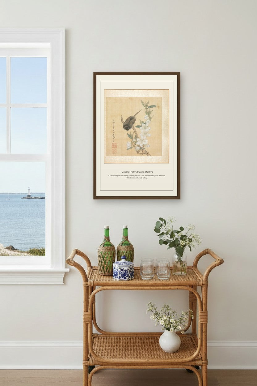 A framed art print featuring a bird on a branch, styled on a wall above a rattan side table with decorative bottles and flowers in a bright room.
