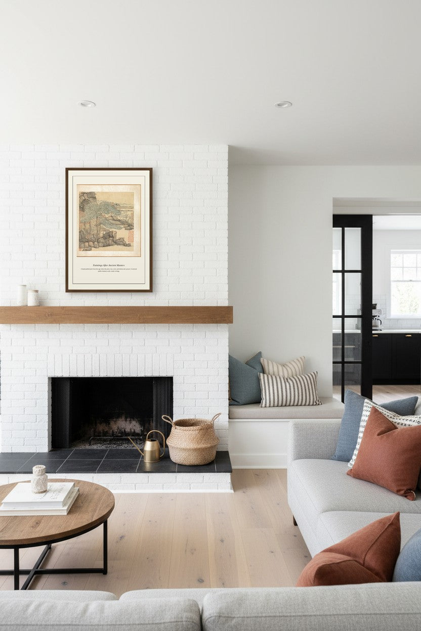 A styled living room interior featuring a white brick fireplace with a wooden mantel, a framed botanical print, a light wood coffee table, and a light gray sofa with rust and blue accent pillows.