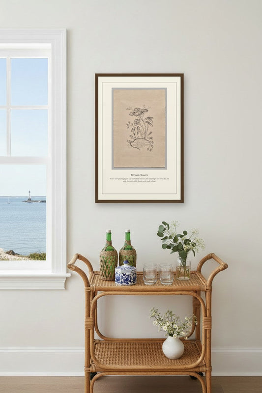 A framed botanical art print hangs on a light gray wall above a rattan bar cart with decorative bottles, glasses, and flowers, next to a window showing the ocean view.