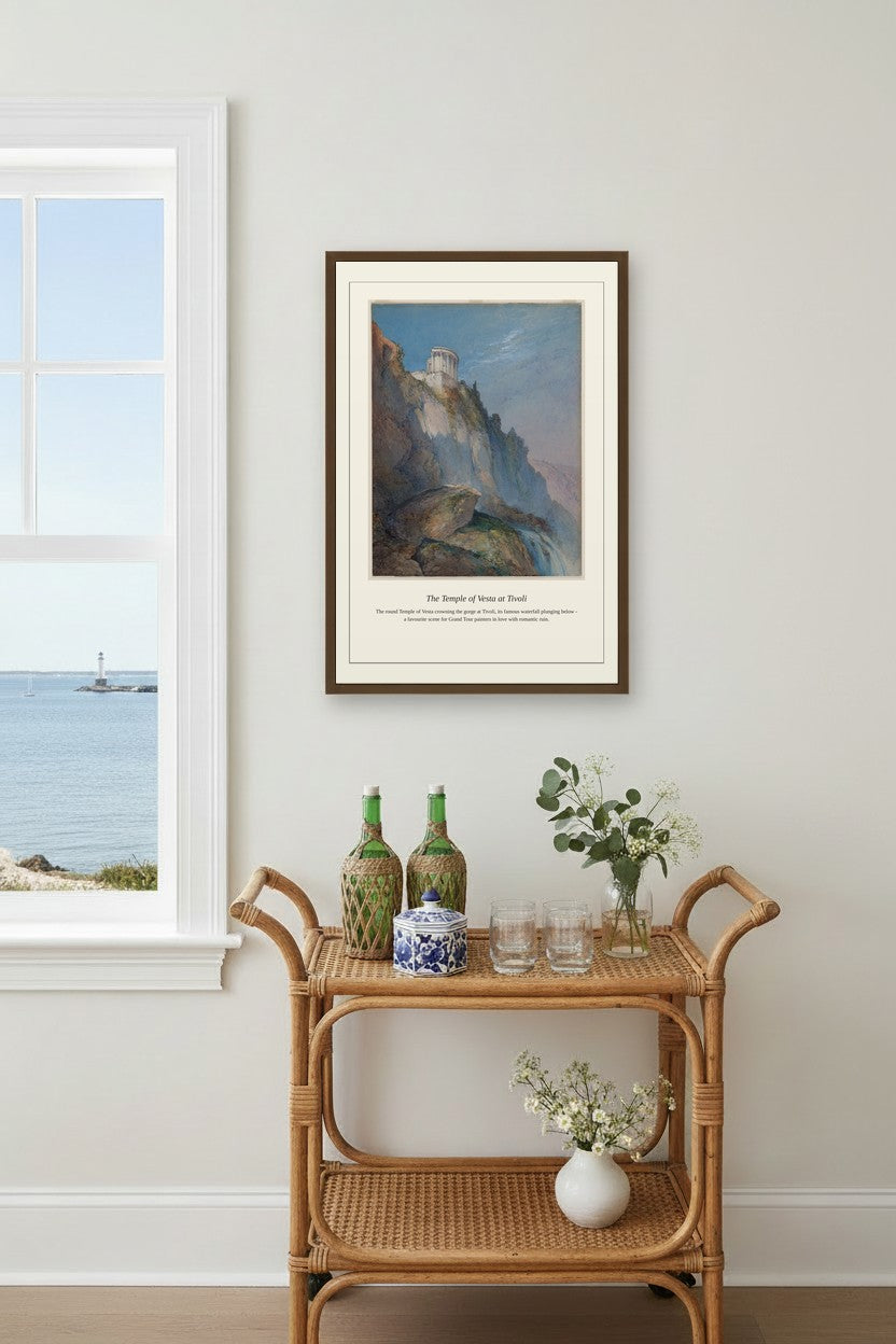 A framed art print titled 'The Cliffs of Dover' hangs on a light gray wall in a styled room setting above a rattan side table with decorative bottles and flowers.